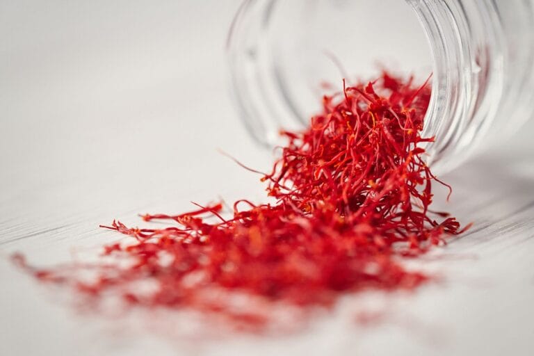Fili di zafferano rosso intenso versati da un barattolo di vetro rovesciato su una superficie di legno chiaro, con un primo piano dettagliato che mette in evidenza la texture e il colore vibrante dello zafferano