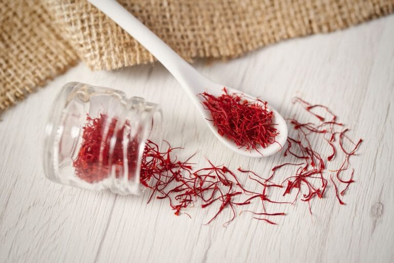 Fili di zafferano rosso intenso sparsi su un tavolo di legno chiaro, con un barattolo di vetro rovesciato e un cucchiaio bianco contenente una piccola quantità di zafferano. Sullo sfondo, un tessuto di juta aggiunge un tocco rustico alla composizione