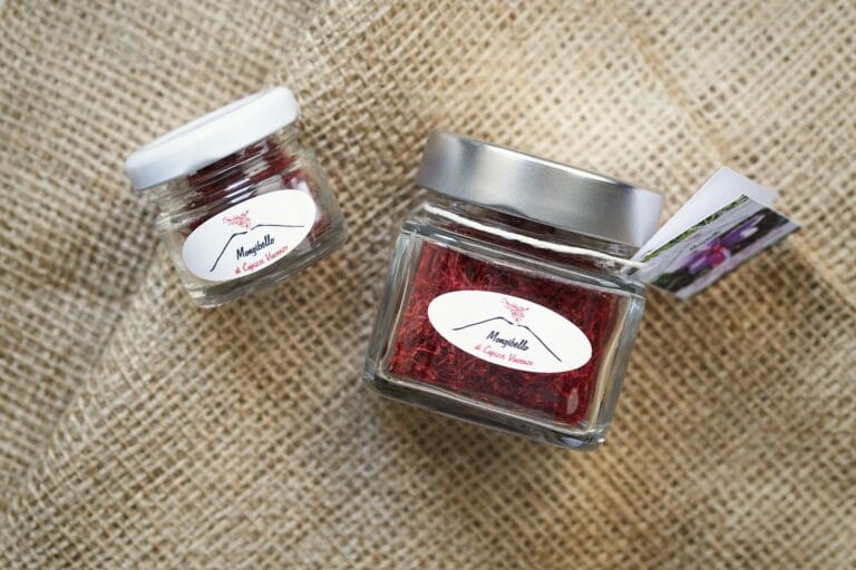 Due barattoli di vetro con coperchio bianco e argento contenenti fili di zafferano rosso intenso, etichettati 'Mongibello di Capizzi Vincenzo', posizionati su un tessuto di juta. Il barattolo più grande ha un'etichetta con un'immagine di un fiore di zafferano allegata con uno spago